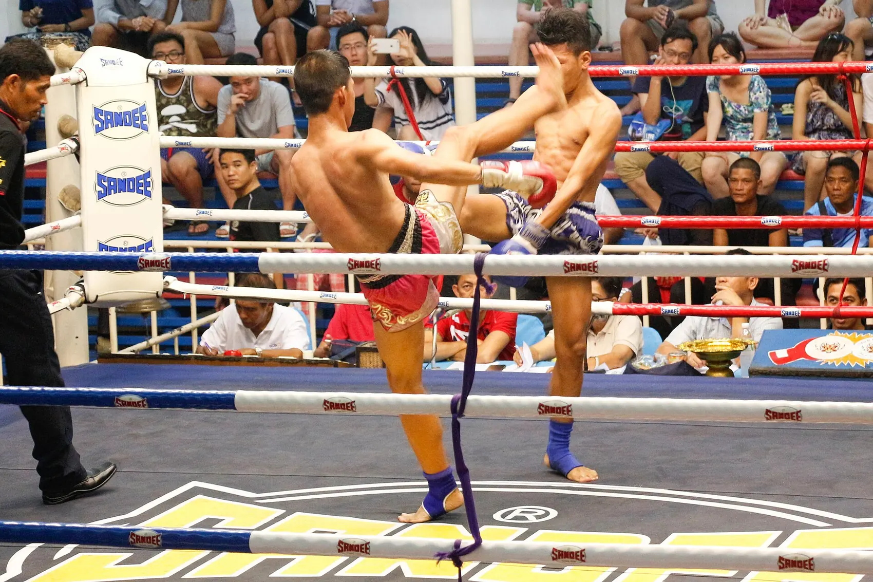Deux boxeurs thaïs échangent un coup de pied et une garde haute sur le ring, devant le public